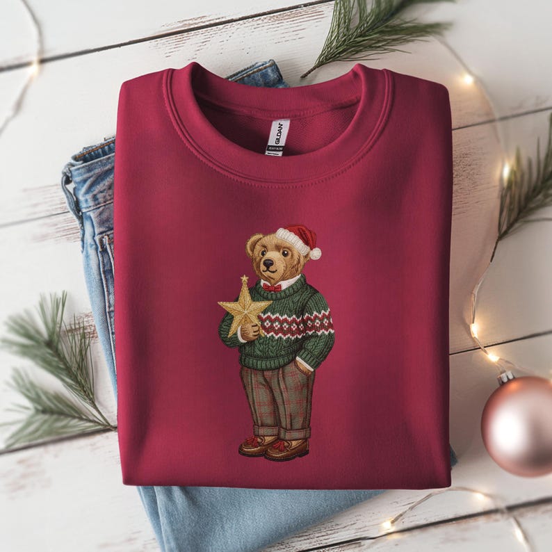 May include: Burgundy crewneck sweatshirt featuring an embroidered bear wearing a Santa hat, green sweater, and plaid pants. The bear holds a gold star. A festive holiday design.