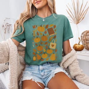 May include: A teal t-shirt featuring a fall-themed graphic. The design includes pumpkins, leaves, a book, a pencil, a candle, and a tote bag with a heart. The shirt is made of a soft material and is suitable for casual wear.