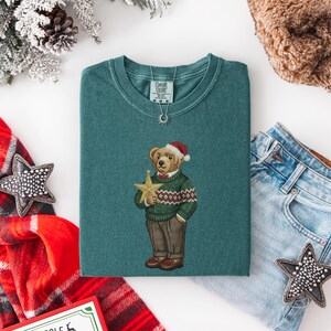 May include: Teal green t-shirt featuring a bear wearing a Santa hat and a green sweater with a red and white pattern. The bear is holding a gold star. The shirt is folded on a white surface, with a pair of jeans and a scarf in the background.