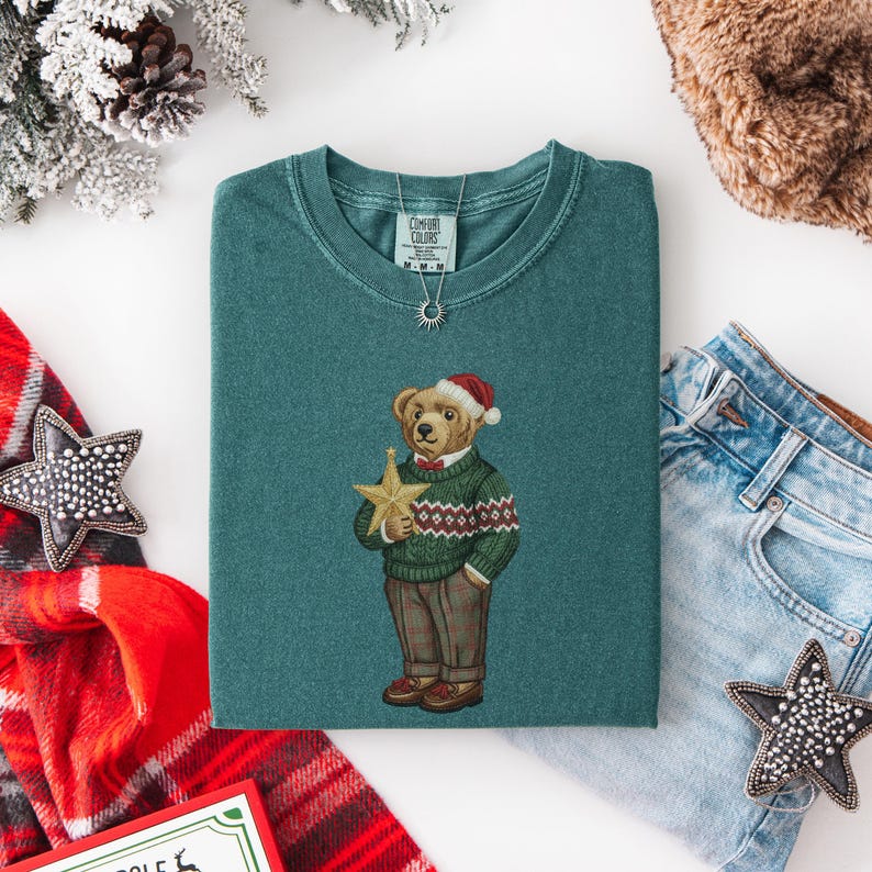 May include: Teal green t-shirt featuring a bear wearing a Santa hat and a green sweater with a red and white pattern. The bear is holding a gold star. The shirt is folded on a white surface, with a pair of jeans and a scarf in the background.