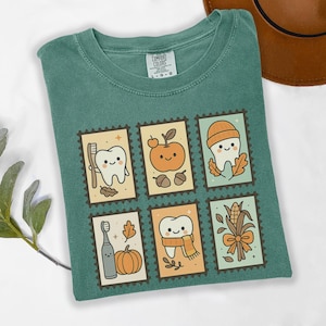 May include: Green Comfort Colors t-shirt featuring six postage stamp-style illustrations. Each stamp depicts a tooth with autumn-themed elements like a toothbrush, pumpkin, apple, and fall leaves. The shirt has a crew neck and short sleeves.