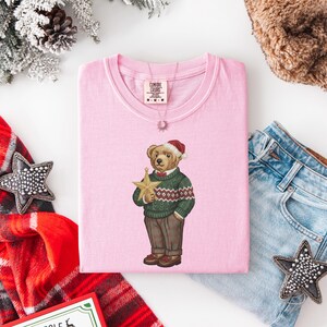 May include: A light pink Comfort Colors t-shirt featuring a bear wearing a Santa hat and a green sweater with a star. The bear is holding a gold star. A silver necklace is on the shirt.