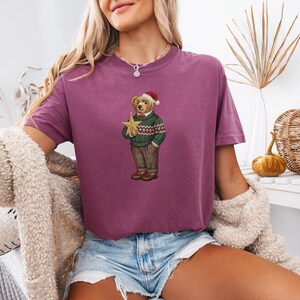 May include: A maroon t-shirt features a Christmas-themed bear illustration. The bear wears a green sweater with a red and white pattern, brown pants, and a Santa hat, holding a gold star. The shirt is paired with denim shorts.
