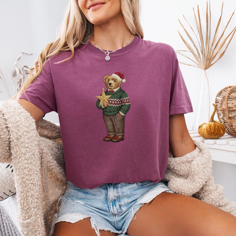 May include: A maroon t-shirt features a Christmas-themed bear illustration. The bear wears a green sweater with a red and white pattern, brown pants, and a Santa hat, holding a gold star. The shirt is paired with denim shorts.