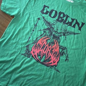 May include: Green t-shirt with a graphic of a goblin, a sword, and the word "GOBLIN" in black. The goblin graphic has red and black accents. The shirt is made of a soft material.