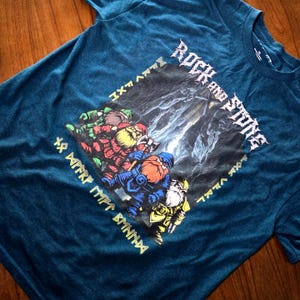 May include: Teal t-shirt featuring a graphic of four cartoon dwarves with axes, set against a dark background. The words "Rock and Stone" are in white, with additional text in a yellow, stylized font. The shirt is made of a soft material.