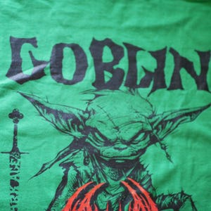 May include: Green t-shirt with a black graphic of a goblin's face and the word "GOBLIN" in a stylized font. A sword graphic is on the left side of the shirt. The goblin has large ears and a stern expression.