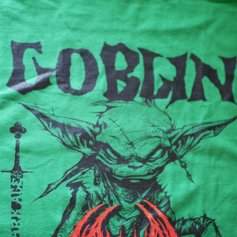 May include: Green t-shirt with a black graphic of a goblin's face and the word "GOBLIN" in a stylized font. A sword graphic is on the left side of the shirt. The goblin has large ears and a stern expression.