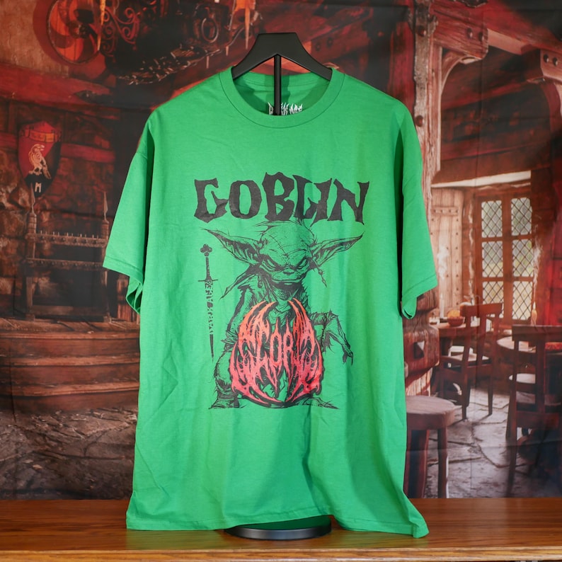 May include: Green t-shirt with a graphic of a goblin holding a sword and a red and pink design. The word "GOBLIN" is printed above the goblin illustration in a black, gothic font. The shirt is displayed on a black hanger.