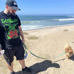 May include: A person wearing a black t-shirt with the word "GOBLIN" in green and a graphic design. The person is wearing a black cap and shorts, standing on a sandy beach with a small dog on a leash.