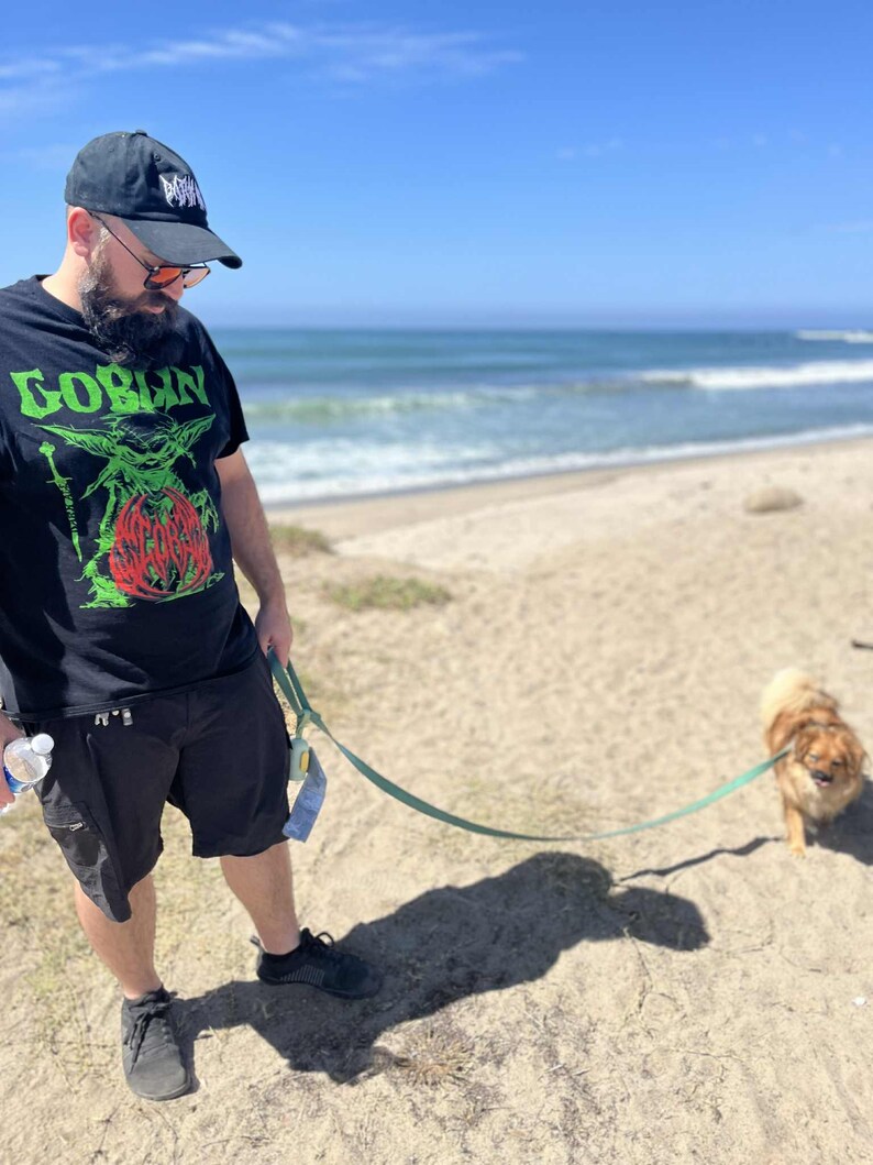 May include: A person wearing a black t-shirt with the word "GOBLIN" in green and a graphic design. The person is wearing a black cap and shorts, standing on a sandy beach with a small dog on a leash.