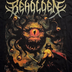 May include: Black t-shirt featuring a detailed illustration. The design includes the word "BEHOLDEN" in a spiky font above a monster with multiple eyes and a warrior with a sword. The artwork is in shades of orange, yellow, and black.