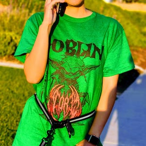 May include: A green t-shirt with the word "GOBLIN" in black block letters above a graphic of a goblin holding a red and orange object. The shirt is worn with a black and white belt.