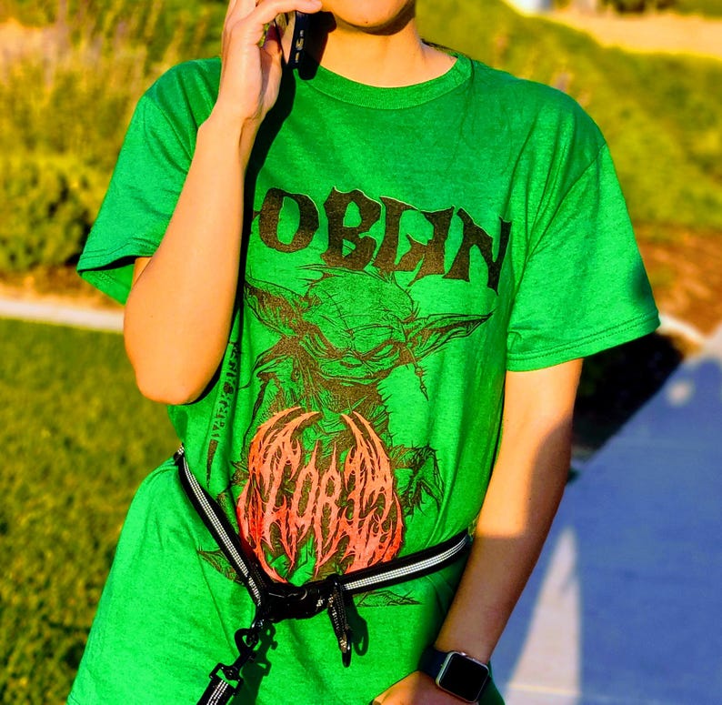 May include: A green t-shirt with the word "GOBLIN" in black block letters above a graphic of a goblin holding a red and orange object. The shirt is worn with a black and white belt.