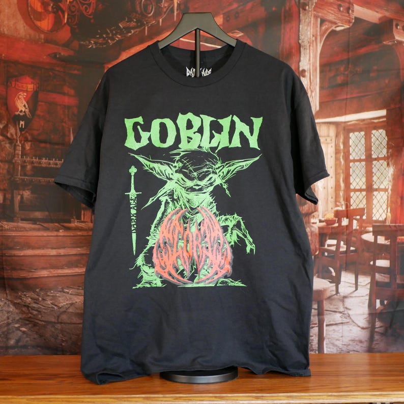 May include: Black t-shirt featuring the word "GOBLIN" in green, above a green and red graphic of a creature with a sword. The shirt is hanging on a black hanger.