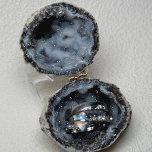 May include: An open geode ring box with a silver ring set inside. The geode is a dark gray and white color with a sparkling interior. The ring set includes a band with small white pearls and a ring with a blue and orange stone.