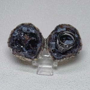 Könnte beinhalten: Eine aufklappbare Geoden-Ringbox mit einem dunklen, kristallinen Inneren in tiefen Violett- und Schwarztönen. Die Geode ist in zwei Hälften geteilt, die durch ein Metallscharnier verbunden sind, und ruht auf einem transparenten Acrylständer.