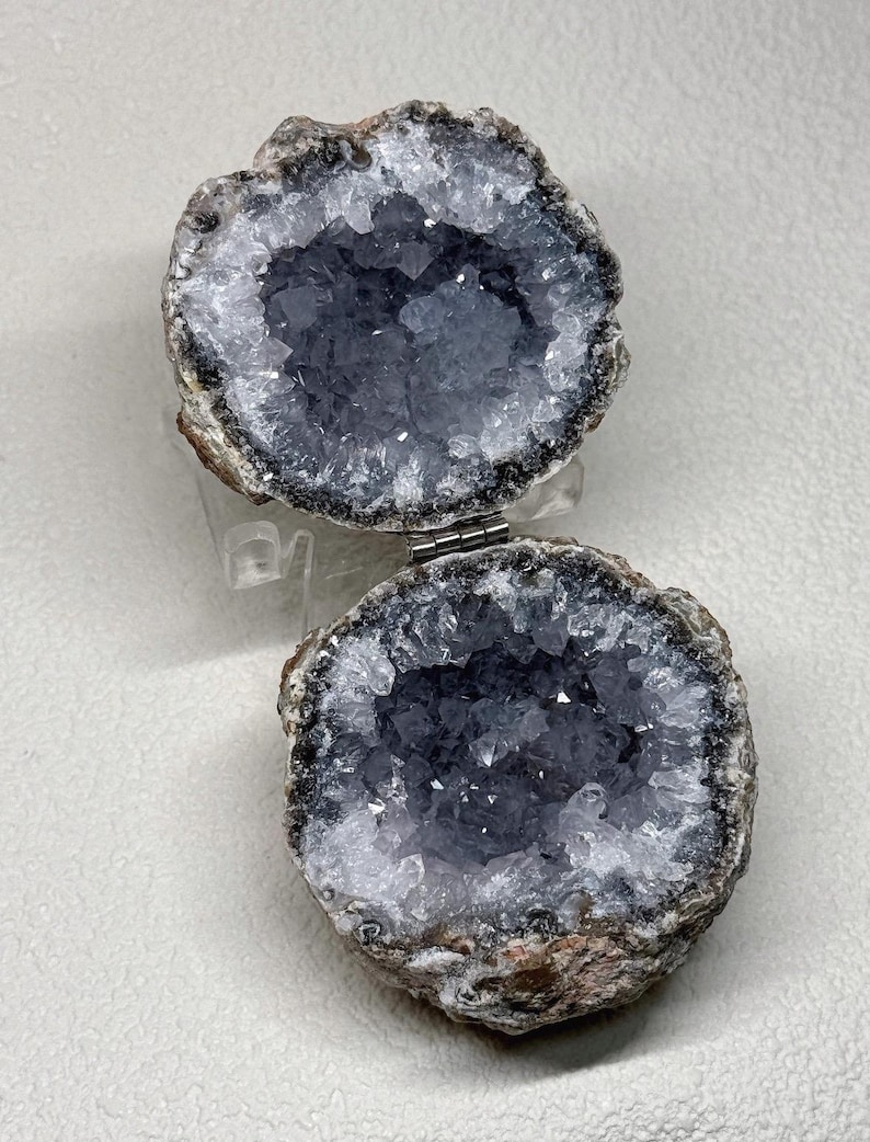 Puede incluir: Una caja geoda con bisagras que presenta dos mitades circulares forradas con cristales de amatista de color p&uacute;rpura intenso. El exterior de la roca es una piedra marr&oacute;n rugosa y texturizada. Una peque&ntilde;a bisagra de metal plateado conecta las dos mitades, lo que permite que la geoda se abra y cierre como un recipiente.