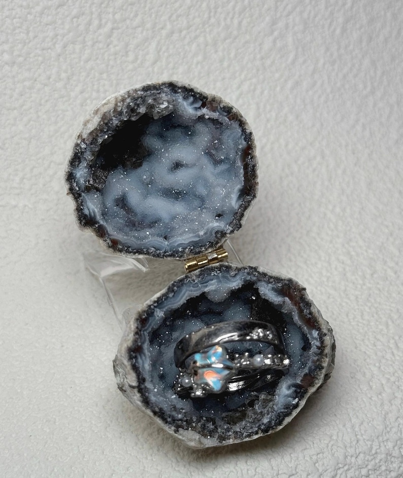 Puede incluir: Un joyero de geoda con bisagras y un interior cristalino oscuro que contiene dos anillos de tono plateado. Un anillo presenta una piedra iridiscente en forma de estrella, mientras que el otro es una banda sencilla con peque&ntilde;as piedras claras.