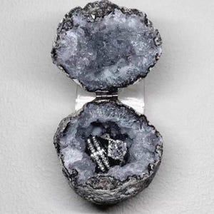 Puede incluir: Un joyero de geoda con un exterior gris natural y rugoso y un interior hueco de cristales brillantes. Las dos mitades est&aacute;n unidas por una bisagra de metal plateado y contienen dos anillos plateados con piedras transparentes facetadas.