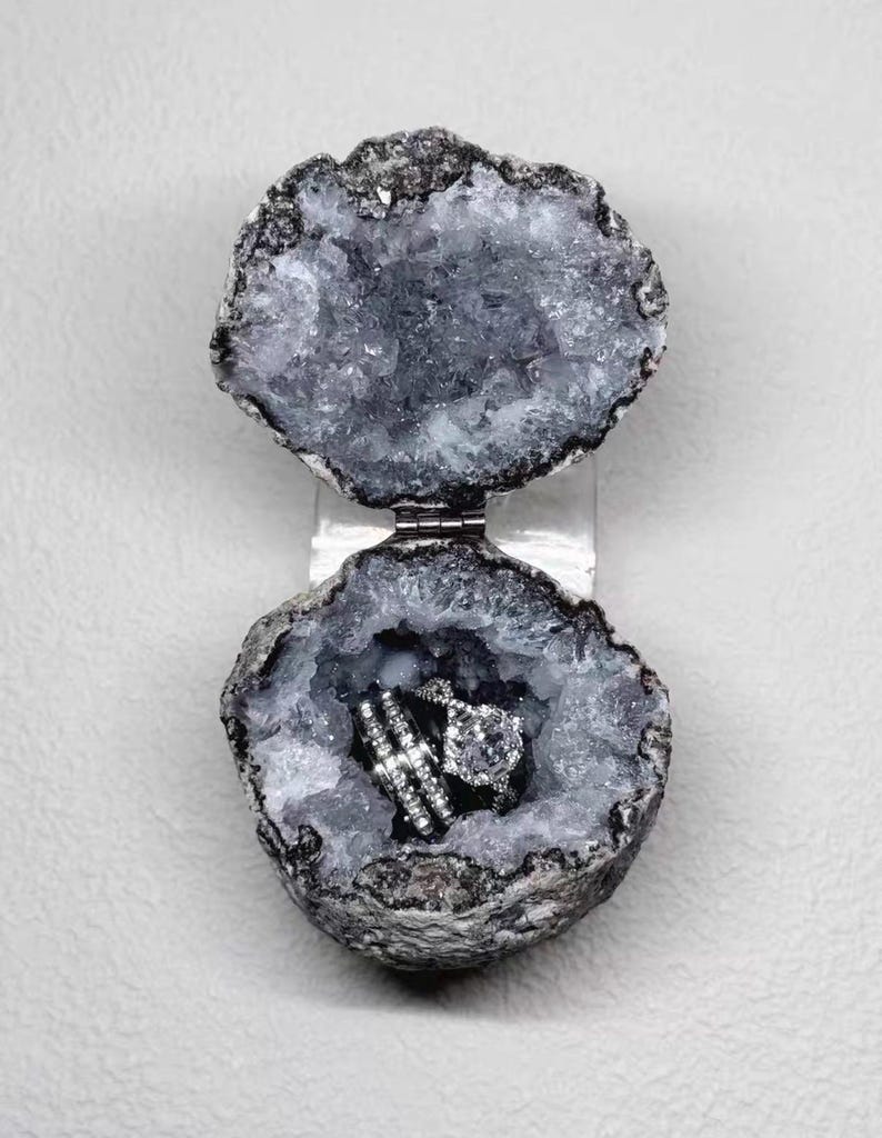 Puede incluir: Un joyero de geoda con un exterior gris natural y rugoso y un interior hueco de cristales brillantes. Las dos mitades est&aacute;n unidas por una bisagra de metal plateado y contienen dos anillos plateados con piedras transparentes facetadas.