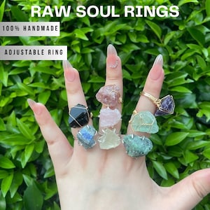 May include: A hand showcases a selection of handmade, adjustable rings. Each ring features a unique, raw gemstone in various colours, including black, blue, pink, green, and purple. The rings are set in gold and silver wire.