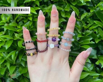 Peut inclure: Une main présente plusieurs mini bagues en cristal. Les bagues sont ornées de pierres précieuses et de perles colorées, dont l'améthyste, la citrine et le quartz clair. Le texte "MINI CRYSTAL RINGS" et "100% HANDMADE" est visible.