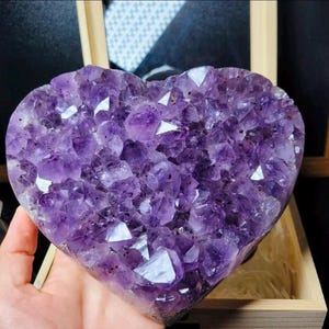 May include: A heart-shaped amethyst crystal cluster. The purple gemstone is composed of many small, sparkling crystals. The heart is held in a wooden box, with the crystals reflecting light.