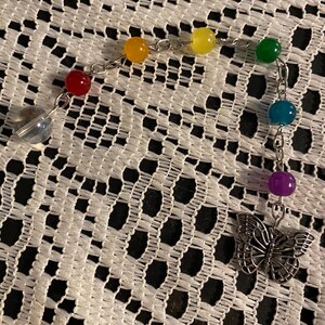 Pode incluir: Uma pulseira de berloques com contas coloridas: vermelho, amarelo, verde, azul e roxo. Apresenta um berloque de borboleta prateada e uma gota de cristal transparente. As contas são enfiadas numa corrente prateada.