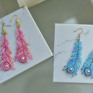 May include: Two pairs of beaded feather earrings. One pair is pink and the other is blue. The earrings have gold-coloured hooks and a pearl-like bead at the bottom. The earrings are displayed on white marble-patterned square boxes.