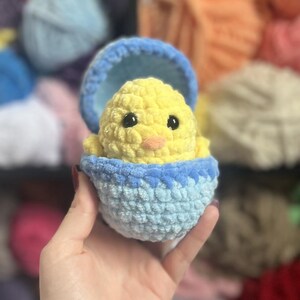 May include: A crocheted chick in a blue and light blue eggshell. The chick is yellow with black eyes and an orange beak. The eggshell is held in a hand, with a blurred background of yarn.