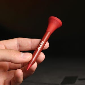 May include: A dark red golf tee with a textured, flared top and a pointed tip. The word "mutée" is printed in white cursive script along the centre of the shaft. The tee features a smooth, matte finish and a bright red accent at the very tip.