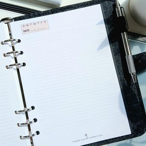 May include: A charcoal gray felt binder with a six-ring metal mechanism holding lined white paper. The top of the page features a box with the text S-M-T-W-T-F-S and DATE, while the bottom includes the text Tangible Tools For Better Care.