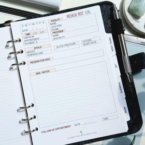 May include: A black ring-bound planner open to a Medical Visit Log page with fields for facility, provider, vitals, and notes. The planner features silver metal rings, a pen holder with a silver pen, and tabbed dividers labeled Dental/Vision, Lab, Diagnostic, and Notes.