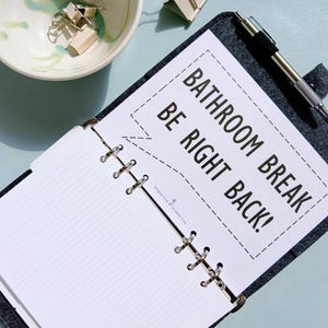 May include: Open gray felt planner with lined paper and a pen. A white paper insert reads "Bathroom Break, Be Right Back!" in black text. A small bowl with gold binder clips sits nearby.