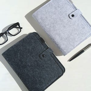 May include: An overhead shot shows two felt journal covers, one gray and one charcoal, with a black pen and black-framed glasses. The journal covers have a button closure. The items are arranged on a white surface.