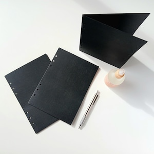 May include: An overhead shot shows black paper sheets, a folded black paper, a pen, and a small bottle with a light pink label. The paper sheets have holes for binding. The pen is silver. The bottle has a wooden top.