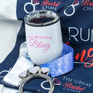 May include: A white stemless wine tumbler with the text "WILL RUN FOR Bling" in pink script, a silver ring with three diamond-shaped accents, and a blue ribbon with a floral design. The background includes a navy blue fabric with the words "The Great Bridal Chase".