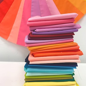 May include: A stack of folded fabric in a variety of colors, including pink, red, orange, yellow, green, blue, and purple. A fan of fabric in a rainbow of colors is in the background. The fabrics are likely for sewing or crafting projects.
