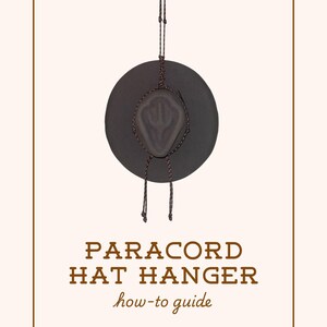 May include: A black hat suspended by a paracord hat hanger. The text "PARACORD HAT HANGER how-to guide" is displayed below the hat, with "by Sky Gennette" underneath. The background is a light beige color.