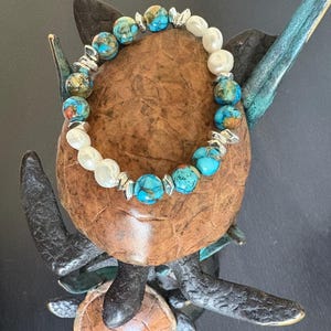 Puede incluir: Un brazalete de cuentas con cuentas turquesas y perlas blancas, intercaladas con detalles plateados. El brazalete se exhibe sobre una superficie marrón texturizada. El conjunto sugiere una pieza de joyería artesanal con una estética decorativa.