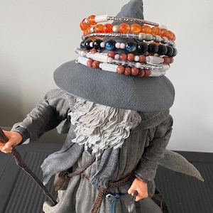May include: A figurine of a wizard in gray robes and a wide-brimmed hat, adorned with multiple beaded bracelets in various colors. The bracelets include orange, black, white, and brown beads. The wizard holds a staff.
