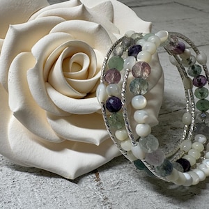 Puede incluir: Una pulsera de múltiples hilos con cuentas blancas, moradas y verdes, apoyada contra una gran rosa color crema. La pulsera presenta un diseño de alambre plateado. Las cuentas son una mezcla de formas redondas y ovaladas.