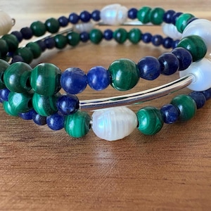 Puede incluir: Una pulsera de múltiples hilos con cuentas de malaquita verde, lapislázuli azul y perlas blancas. Las cuentas están ensartadas en un alambre plateado. La pulsera se exhibe sobre una superficie de madera.