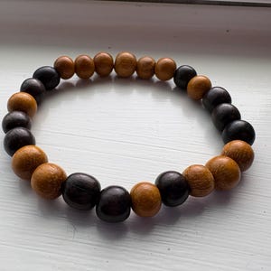 Puede incluir: Una pulsera de cuentas de madera con una mezcla de cuentas marrón oscuro y marrón claro. La pulsera es circular y parece estar ensartada en un cordón elástico. Las cuentas son lisas y redondas, creando una estética natural y terrosa.
