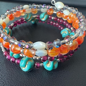 Può includere: Un braccialetto avvolgente a più fili con un mix di perline colorate. Il braccialetto include perline arancioni, bianche, viola e turchesi, insieme a perline trasparenti e sfaccettate. Le perline sono infilate su un filo color argento, creando un design a spirale.