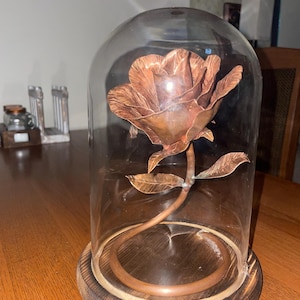 May include: A copper-colored rose sculpture with leaves and stem, encased in a clear glass cloche. The cloche sits on a round, wooden base. The rose is detailed and appears to be made of metal. The overall aesthetic is decorative.