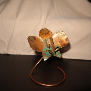 Könnte beinhalten: Eine handgefertigte Kupferblumenskulptur mit gebogenem Stiel. Die Blütenblätter zeigen eine Mischung aus Bronze- und Grünspan-Farbtönen. Die Skulptur ist etwa 15 cm hoch.