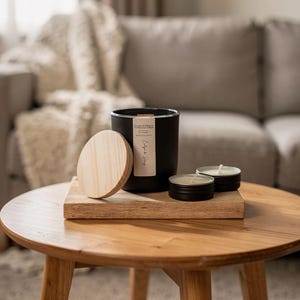 Peut inclure: Une bougie noire dans un récipient cylindrique avec un couvercle en bois, posée sur un plateau en bois. Deux petites bougies rondes sont à côté. Le récipient de la bougie a une étiquette avec du texte. Sur une table ronde en bois.