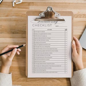 Peut inclure: Un presse-papiers avec une "Liste de contrôle de désencombrement" imprimée sur une feuille de papier blanche. La liste de contrôle comprend diverses tâches de désencombrement. Une main tient un crayon, prêt à cocher la liste. Le presse-papiers est sur une surface en bois.